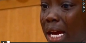 Still from video, young person wtih dark skin, looking past the camera in tears, with an expression of grief on their face