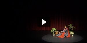 Person in an orange and purple robe sits on a stage near a plant, in front of a red curtain, reciting poetry.